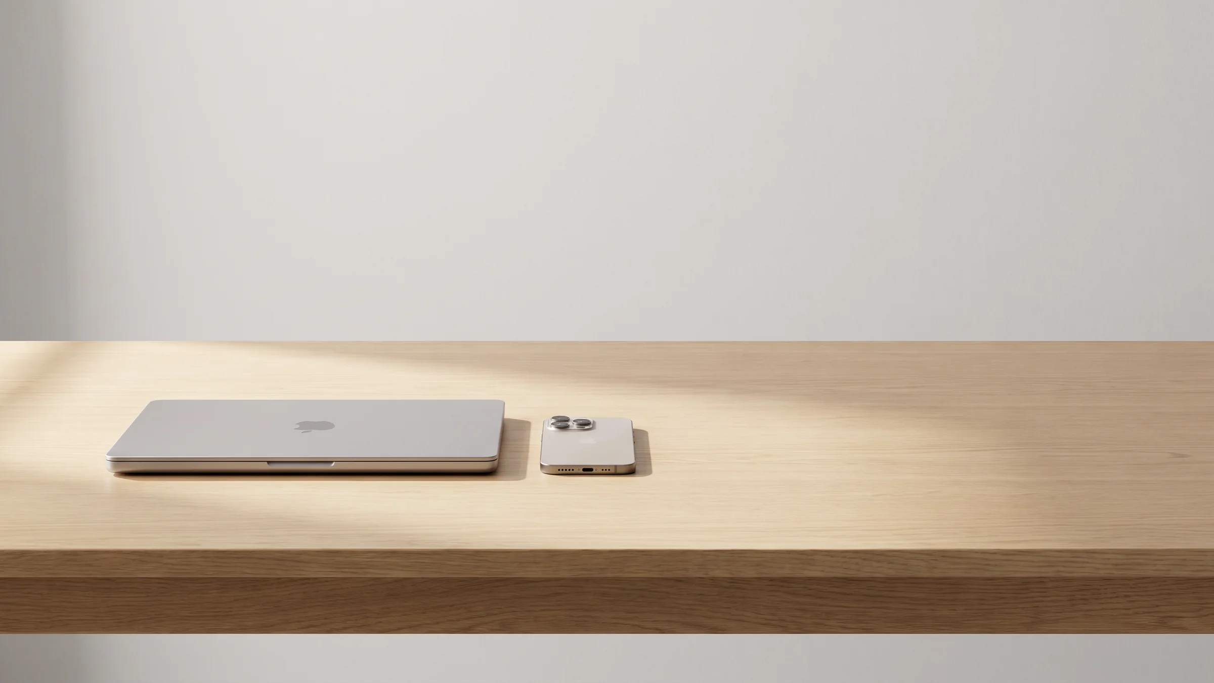A closed MacBook Pro and an iPhone face down on a light oak desk in soft daylight